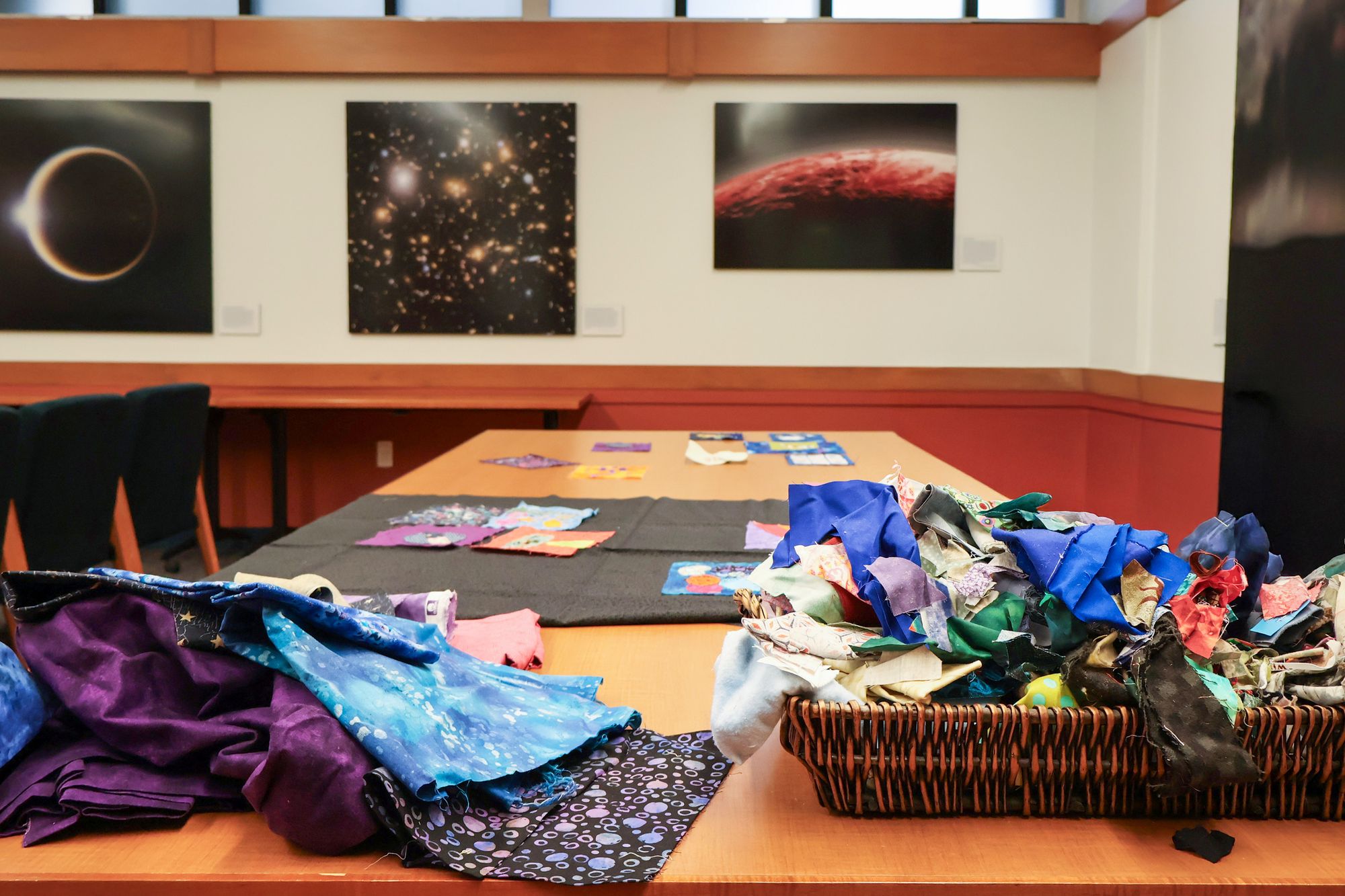 A long wooden table covered with fabric scraps, baskets of materials, and quilt squares on display, set against a backdrop of space-themed wall art.