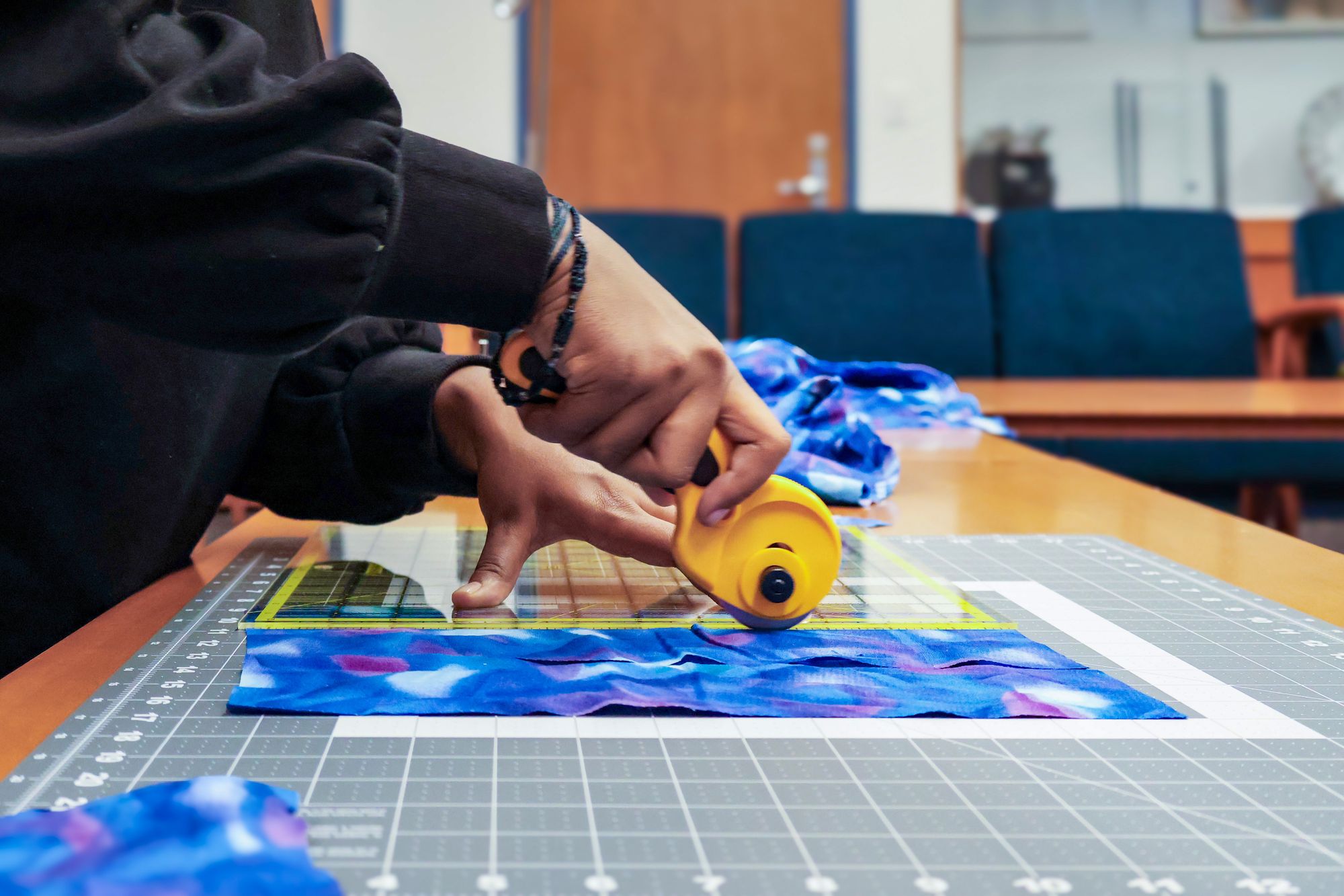Close-up of hands holding a rotary cutter, slicing through blue patterned fabric against a yellow ruler and cutting mat.