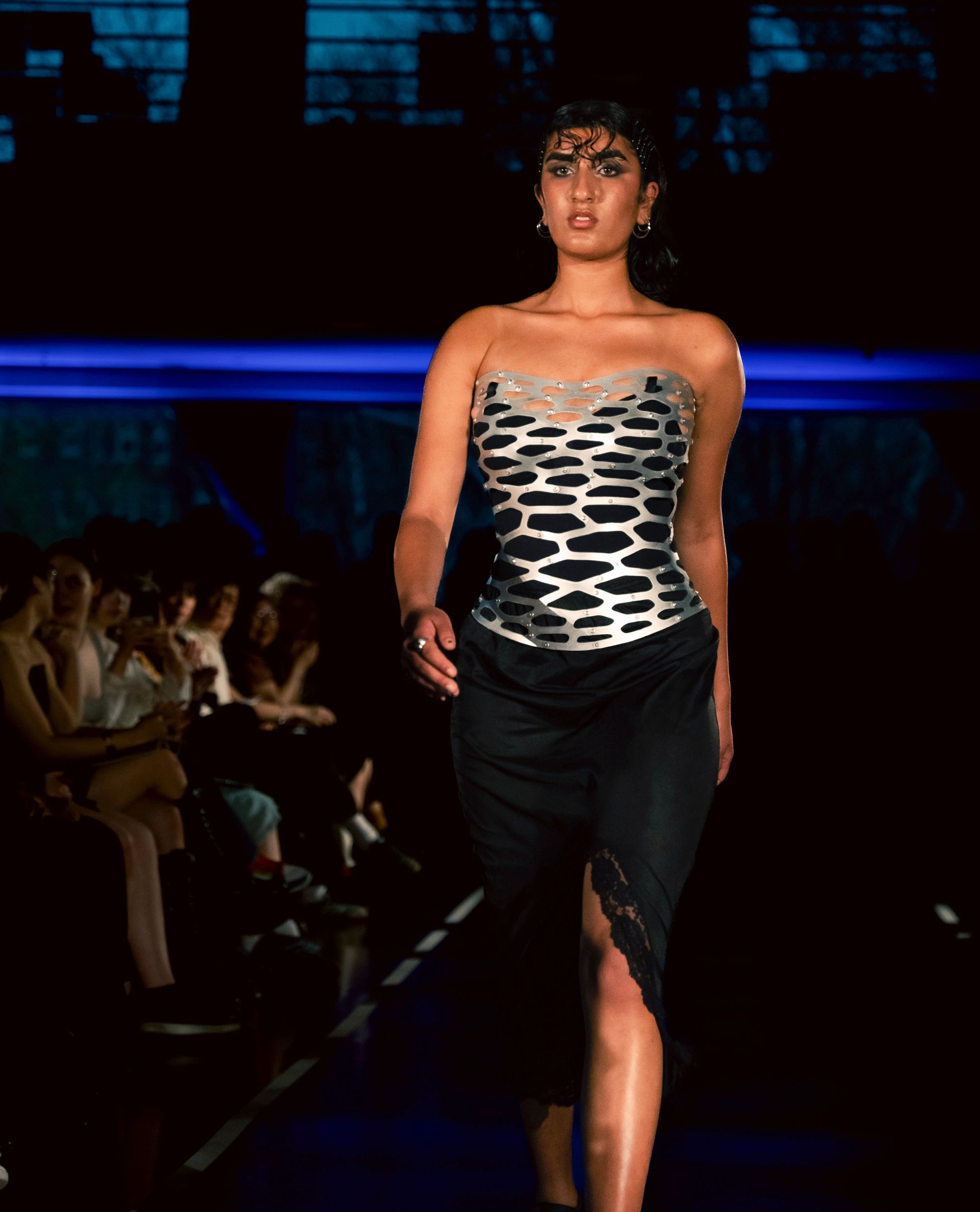 Model walks the runway in a striking strapless metallic corset composed of interlocking, perforated segments forming an organic, web-like pattern. The corset is paired with a black midi skirt featuring a high slit edged in lace. The model wears hoop earrings and a slick, wet-look hairstyle under dramatic lighting.