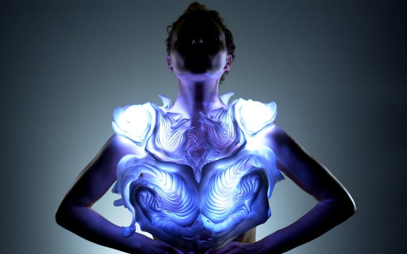 A person stands with arms on their hips, wearing a sculptural, glowing garment that emits blue and white light. The piece has layered, organic patterns resembling rippling waves or carved topography. The wearer’s face is shadowed, creating a dramatic focus on the illuminated design across the chest and shoulders.