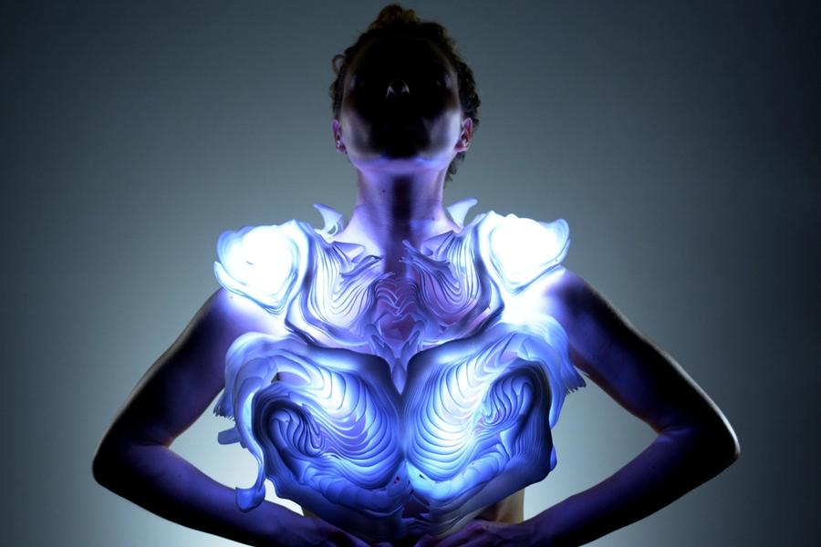 A person stands with arms on their hips, wearing a sculptural, glowing garment that emits blue and white light. The piece has layered, organic patterns resembling rippling waves or carved topography. The wearer’s face is shadowed, creating a dramatic focus on the illuminated design across the chest and shoulders.