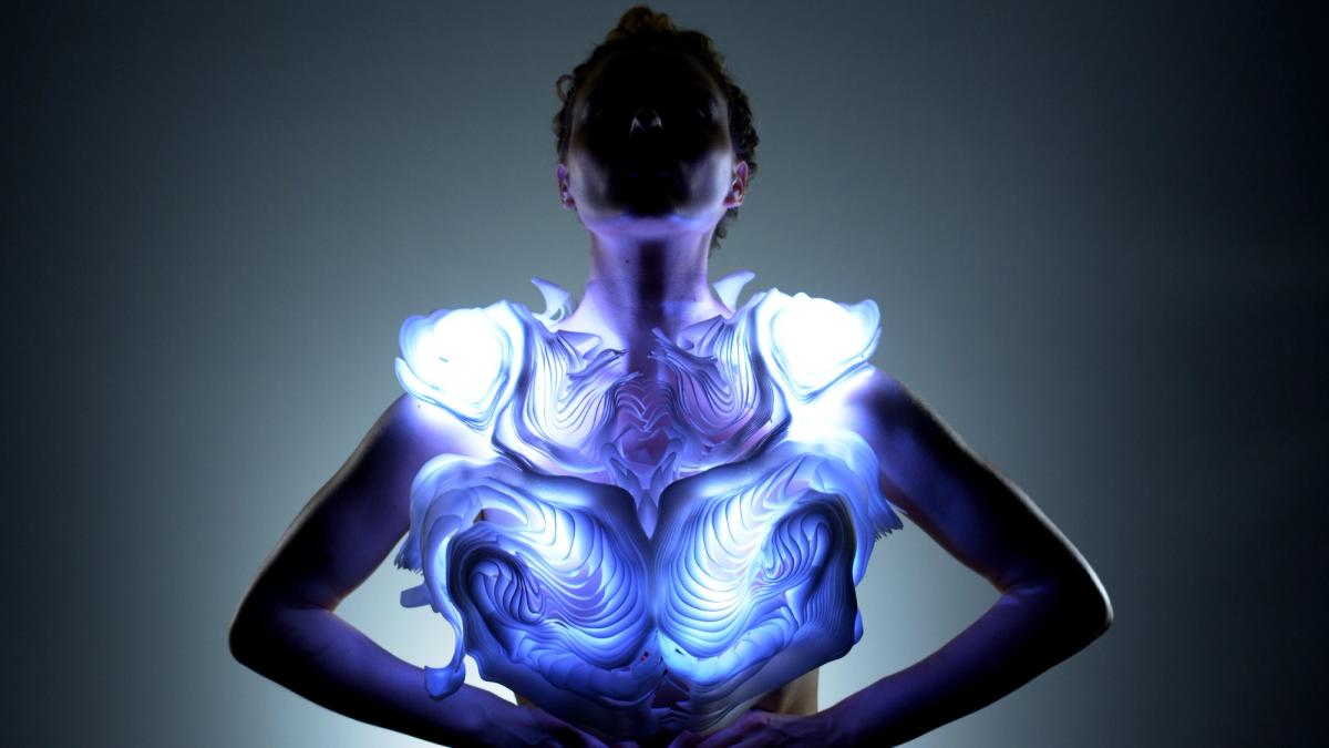A person stands with arms on their hips, wearing a sculptural, glowing garment that emits blue and white light. The piece has layered, organic patterns resembling rippling waves or carved topography. The wearer’s face is shadowed, creating a dramatic focus on the illuminated design across the chest and shoulders.