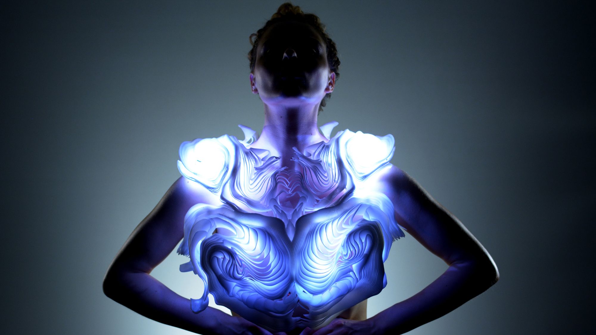 A person stands with arms on their hips, wearing a sculptural, glowing garment that emits blue and white light. The piece has layered, organic patterns resembling rippling waves or carved topography. The wearer’s face is shadowed, creating a dramatic focus on the illuminated design across the chest and shoulders.