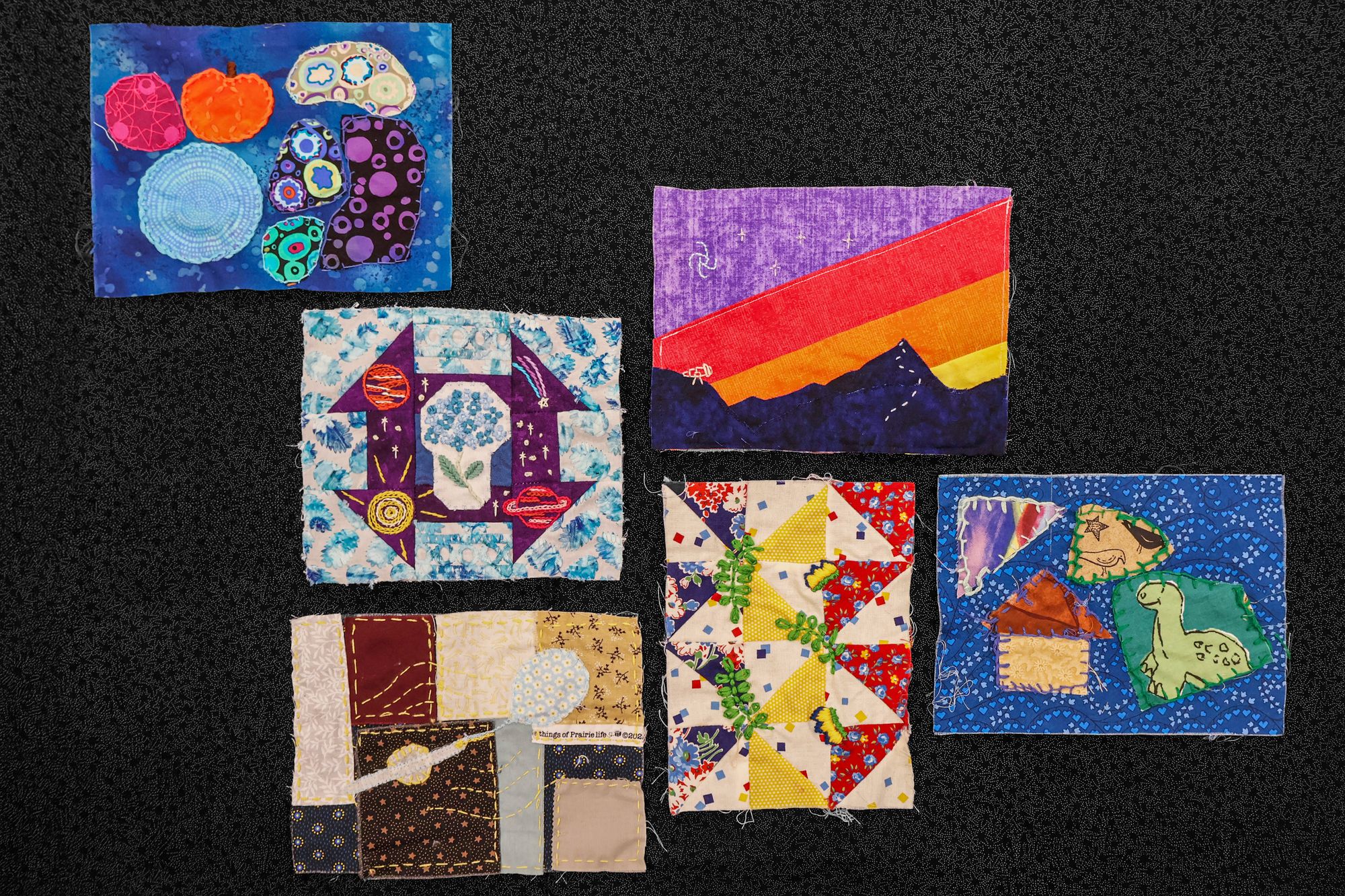 Six quilt squares displayed together, each with colorful fabric and embroidery: designs include planets, flowers, mountains, geometric patterns, and playful figures.