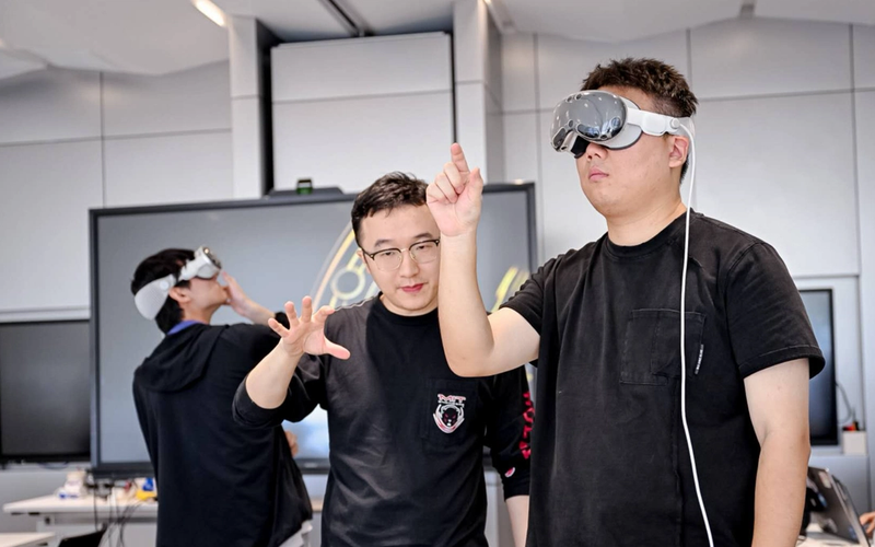 Two students in a classroom setting use virtual reality headsets. One student wears a black shirt and headset while extending his arm and pointing forward. Another student, standing beside him in glasses and a black shirt with an MIT logo, gestures with his hand as if guiding him. In the background, a third student also wears a headset and gestures with his hand.
