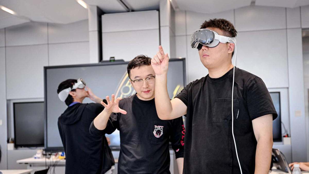 Two students in a classroom setting use virtual reality headsets. One student wears a black shirt and headset while extending his arm and pointing forward. Another student, standing beside him in glasses and a black shirt with an MIT logo, gestures with his hand as if guiding him. In the background, a third student also wears a headset and gestures with his hand.