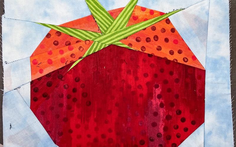 Paper-pieced quilt block featuring a bright red tomato with dark spots, an orange top, and green striped leaves, set against a pale blue sky-like background. The tomato is made from various patterned fabrics assembled into angular shapes.