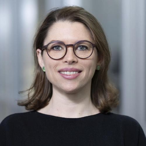 A smiling woman with light brown hair and glasses, wearing a black top, poses for a headshot against a blurred indoor background with glass and metal elements.