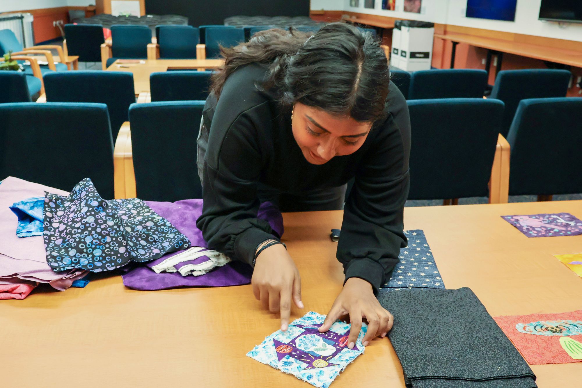 A woman points to a colorful quilt square decorated with stitched shapes while leaning over a table covered with fabric pieces.