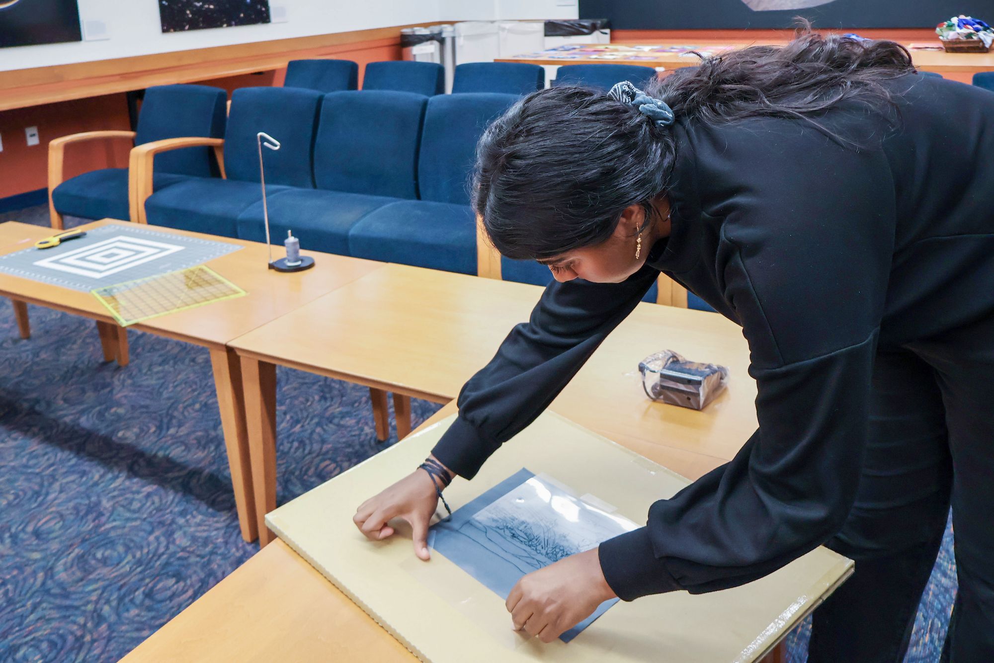 A woman leans over a table, carefully pressing a sheet with a line drawing onto a board. Chairs and craft materials are visible in the background.