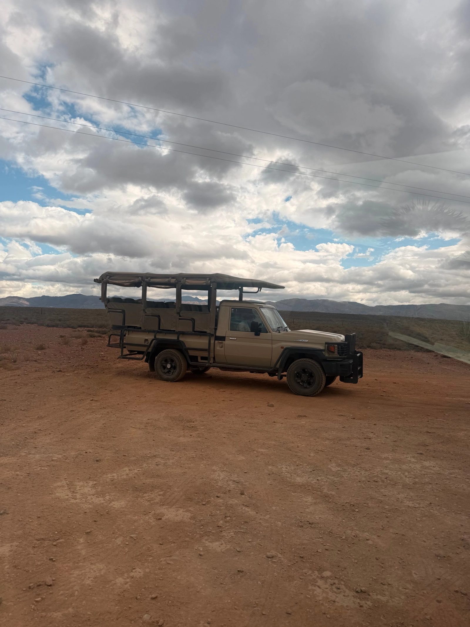 Open game-drive vehicle parked on red Karoo earth under a broken cloudy sky at Inverdoorn