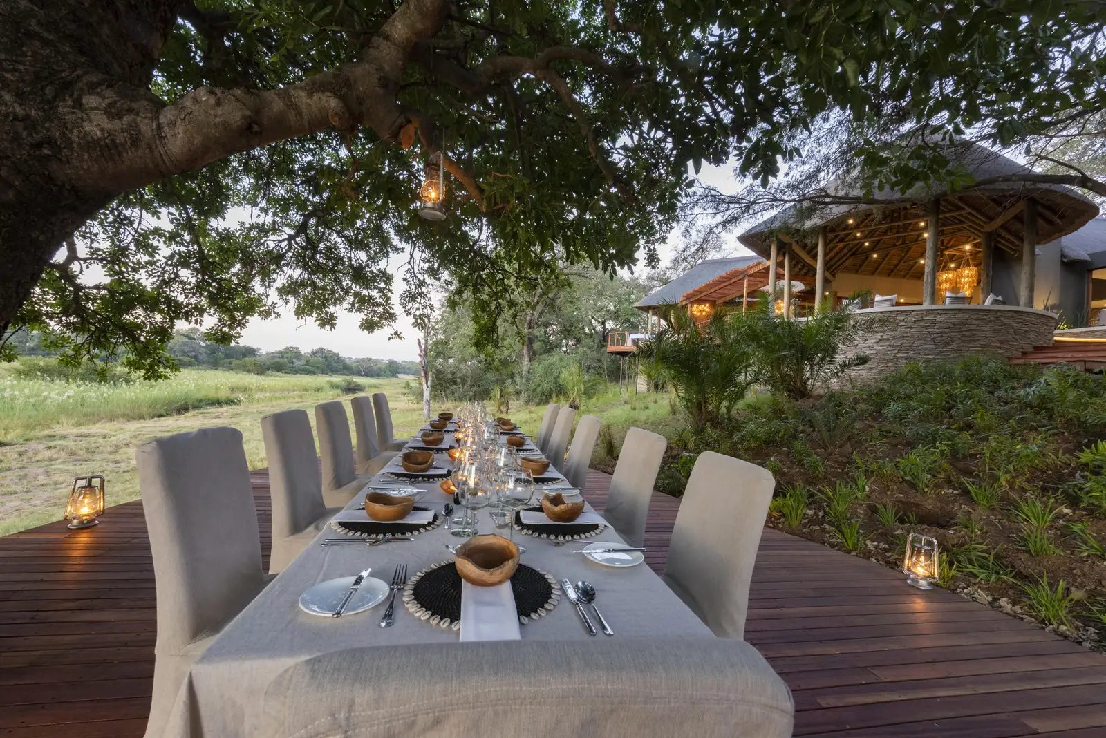 Long dinner table set on the Dulini River Lodge deck under an ebony tree with the thatched lodge behind