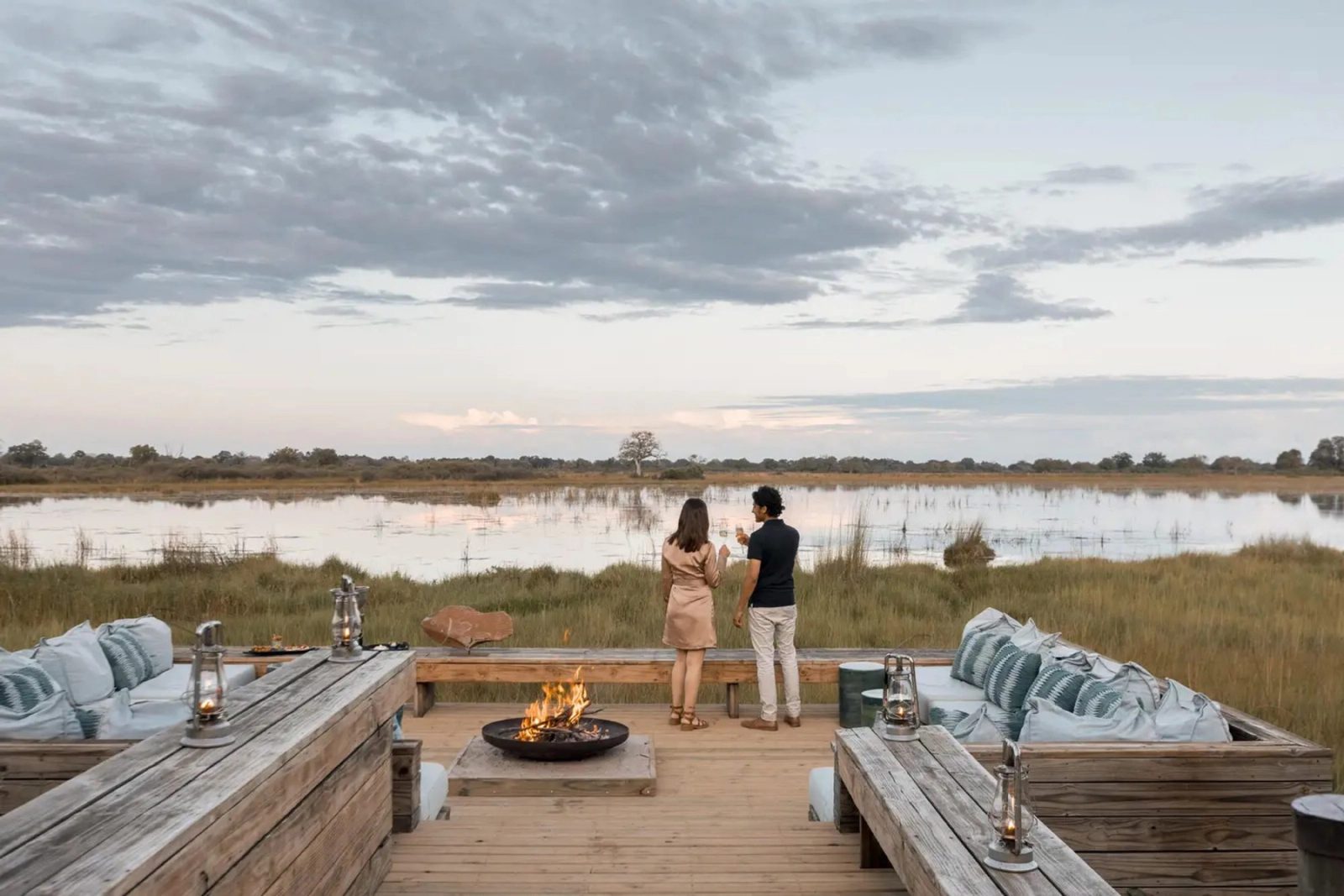 A couple stand with drinks at a fire-pit deck overlooking a floodplain at dusk