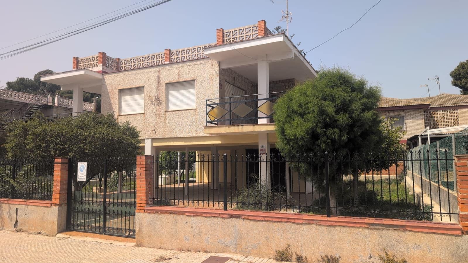 Front of a detached two-storey villa with textured render, closed white roller shutters, a balcony with diamond-pattern railing, a breeze-block roof parapet between brick piers and an open columned porch, behind a low wall with black iron railings and mature trees.