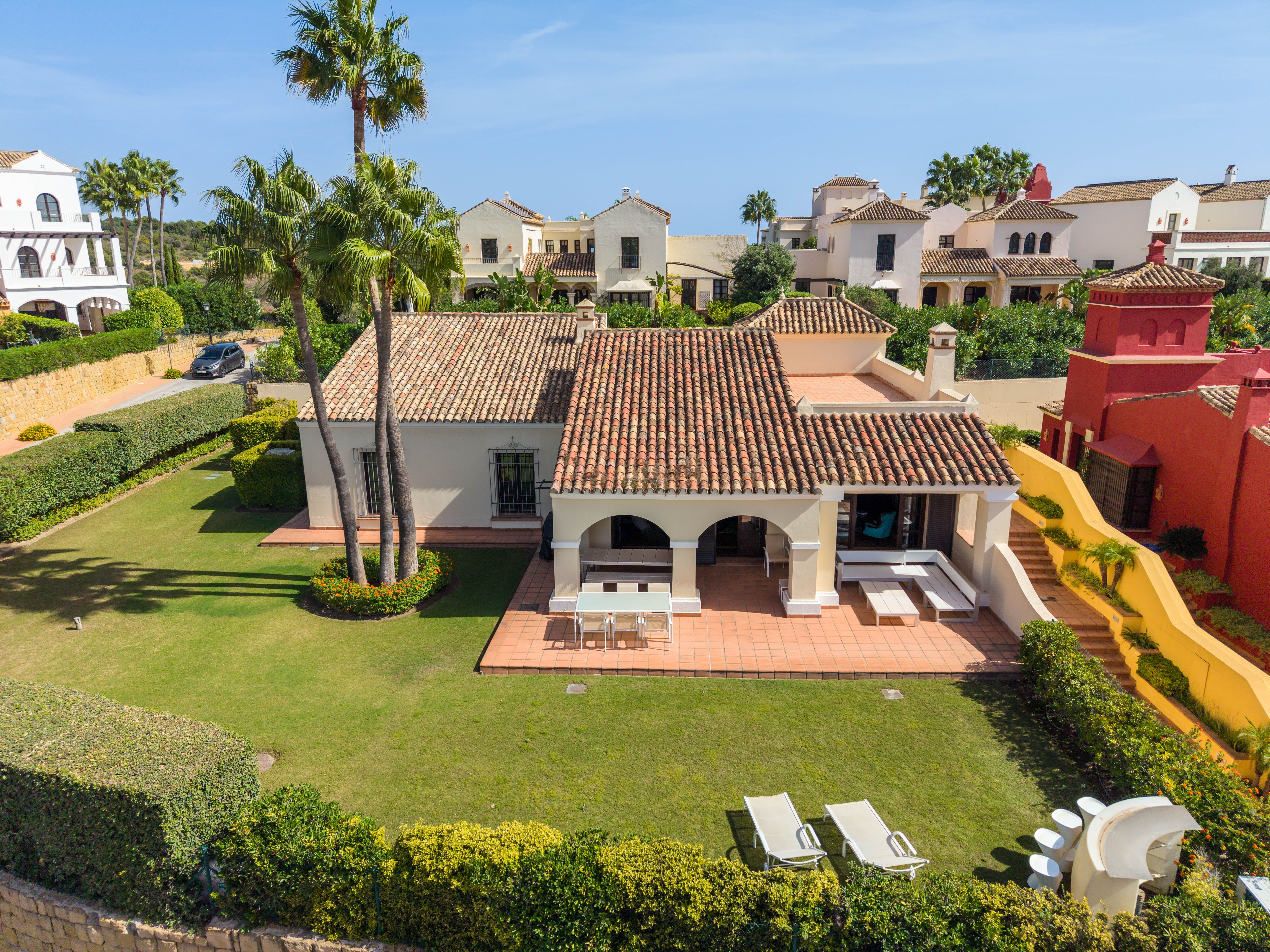Aerial view of an Andalusian-style villa in Los Cortijos de La Reserva, Sotogrande, with tiled roofs, an arched covered terrace, tall palms and a private lawn garden