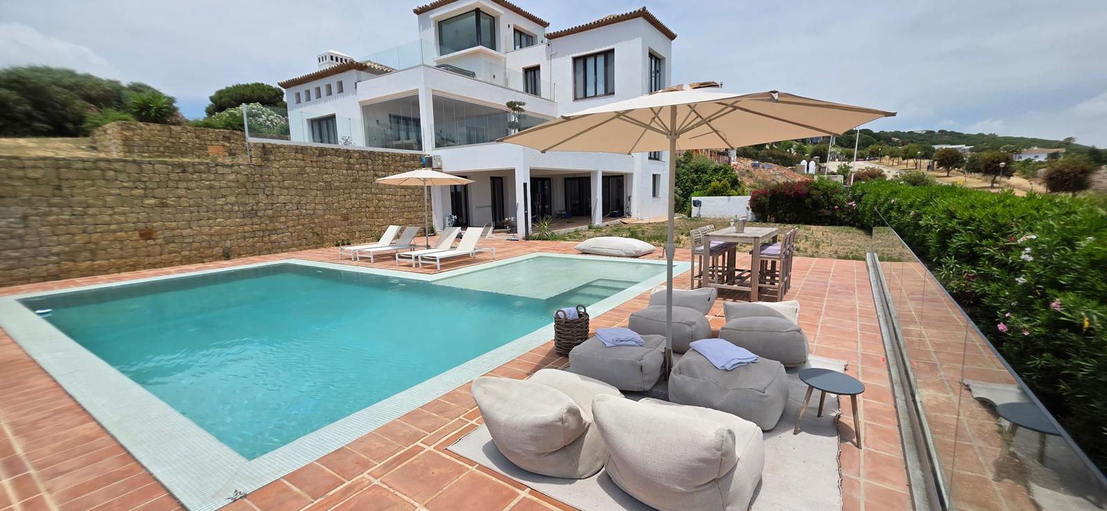 Multi-level white villa with terracotta roofs rising above an L-shaped swimming pool, lounge terrace with poufs and parasols, and a natural stone wall