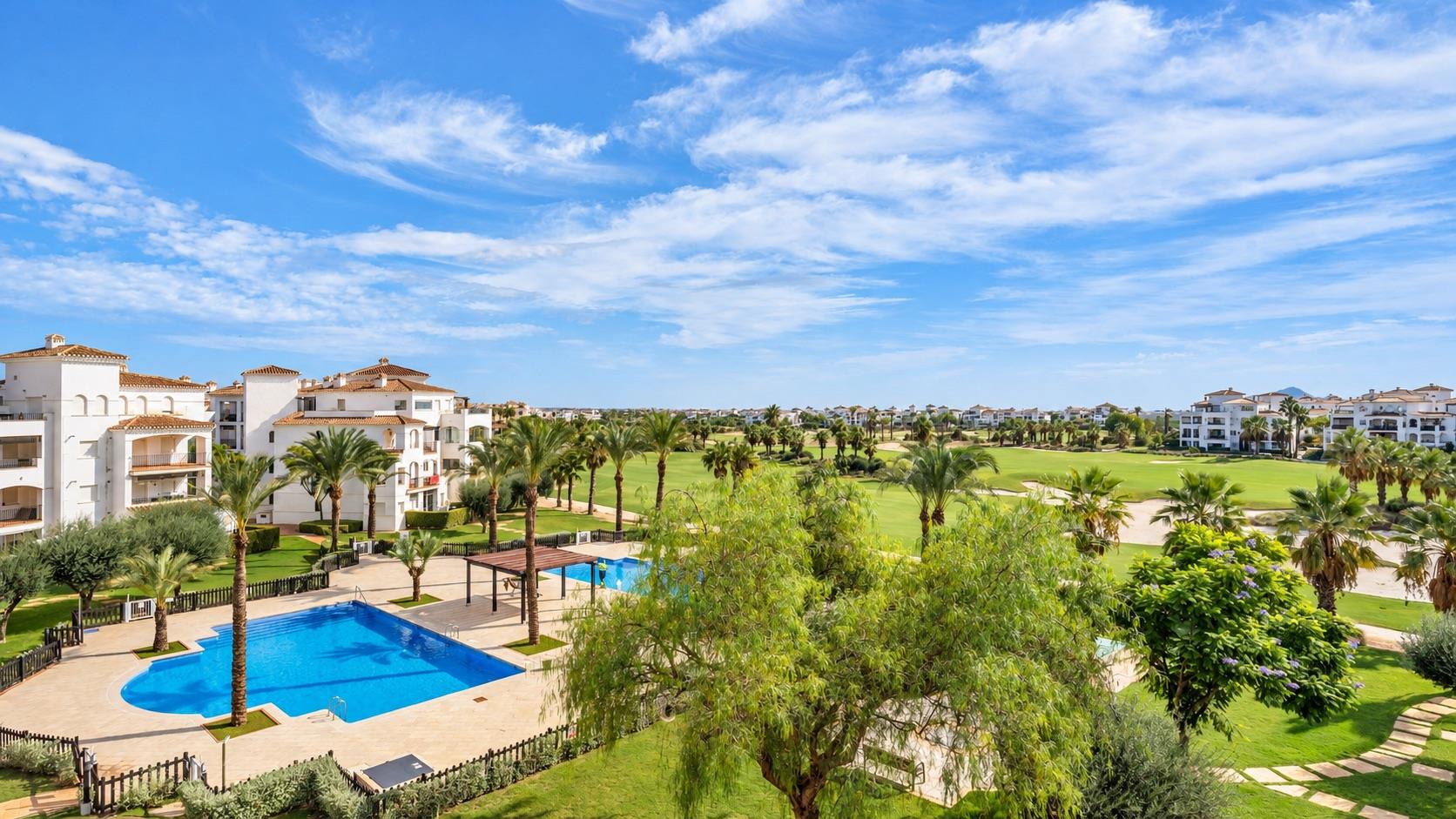 Elevated view over a resort with a large kidney-shaped communal pool, a second pool beside a timber pergola, palm trees, white terracotta-roofed apartment blocks and green golf fairways beyond.
