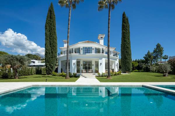 Villa Karina exterior with private pool, white villa facade, landscaped garden, cypress trees and mountain backdrop in Nueva Andalucia.