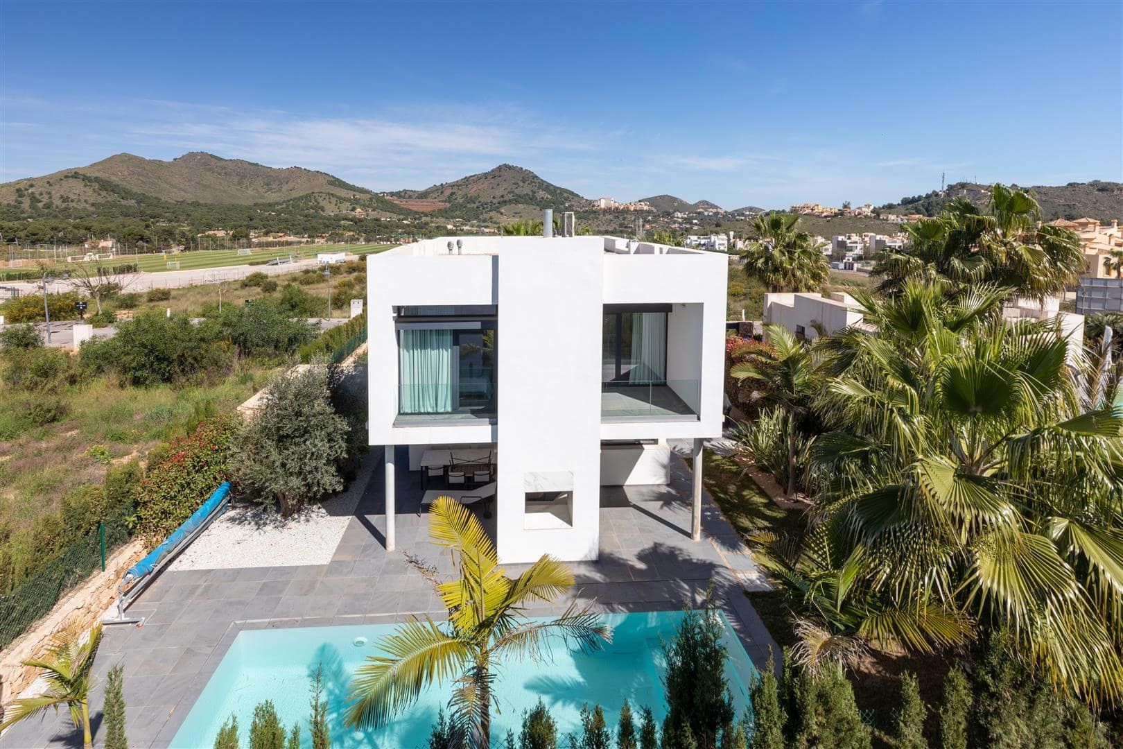Aerial daytime view of a contemporary white villa, swimming pool, palms and surrounding hills