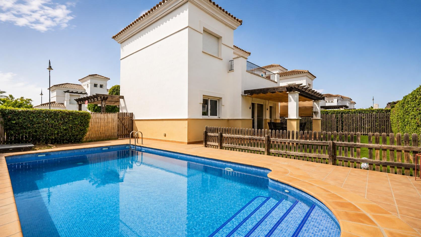 Private-garden swimming pool with blue mosaic lining, Roman corner steps and terracotta coping, in front of a white two-storey villa with a tower, upper balcony and timber-beamed covered terrace, screened by hedges and reed fencing.