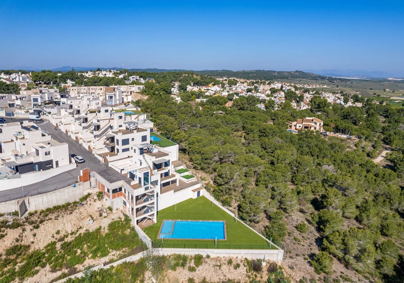 Day aerial view of white residential buildings beside wooded land with a rectangular swimming pool in the foreground