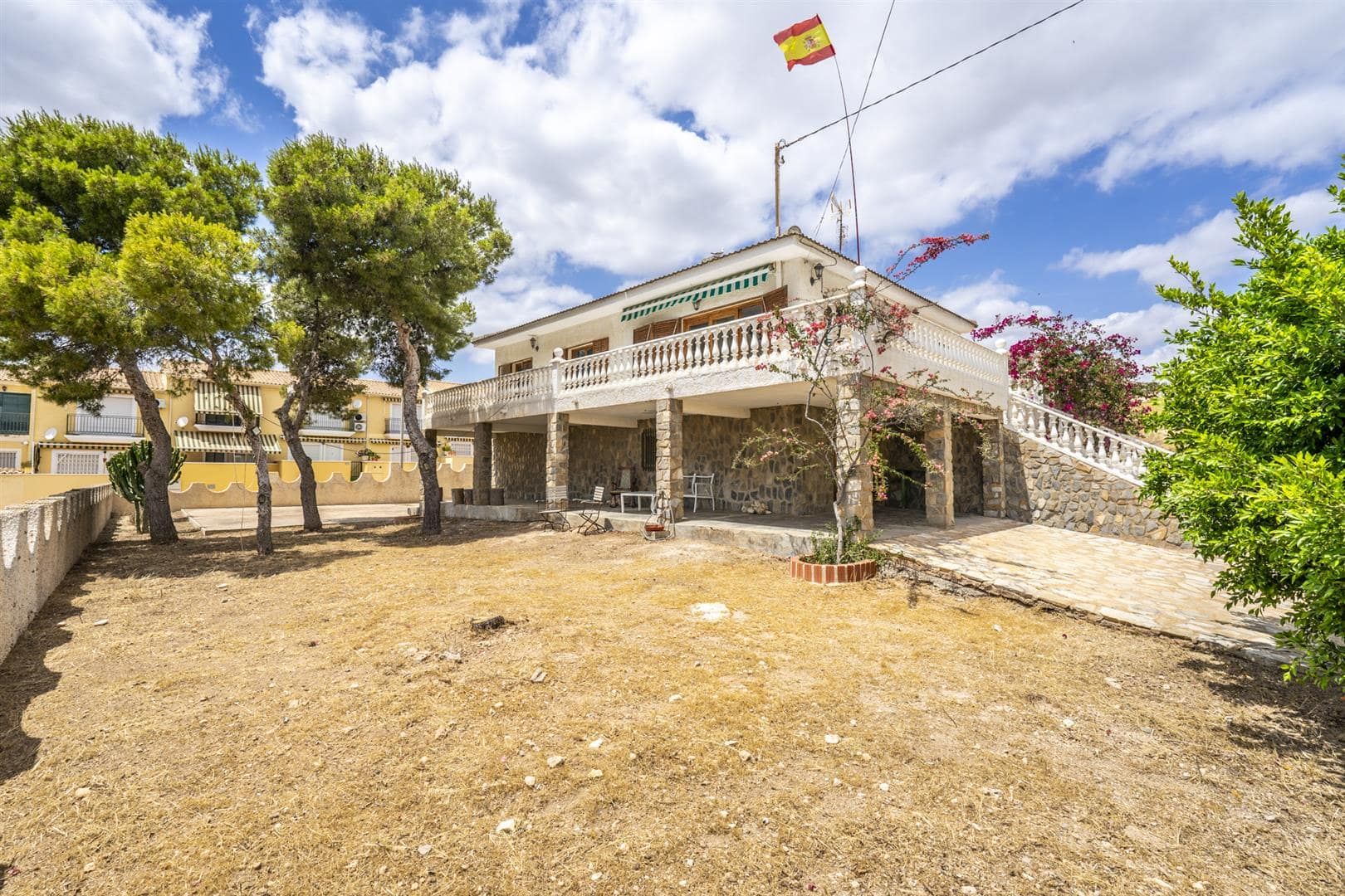 Wide front view of the detached villa, stone lower level, upper terrace and mature trees.