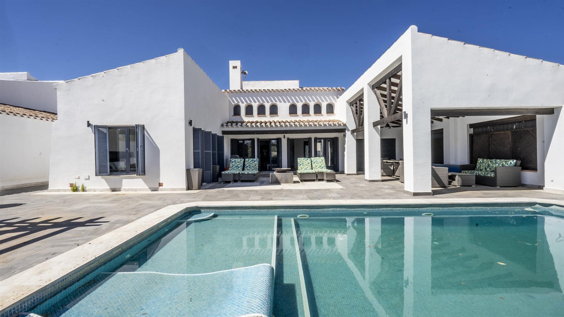 White villa arranged around a swimming pool and paved terrace under a clear blue sky.