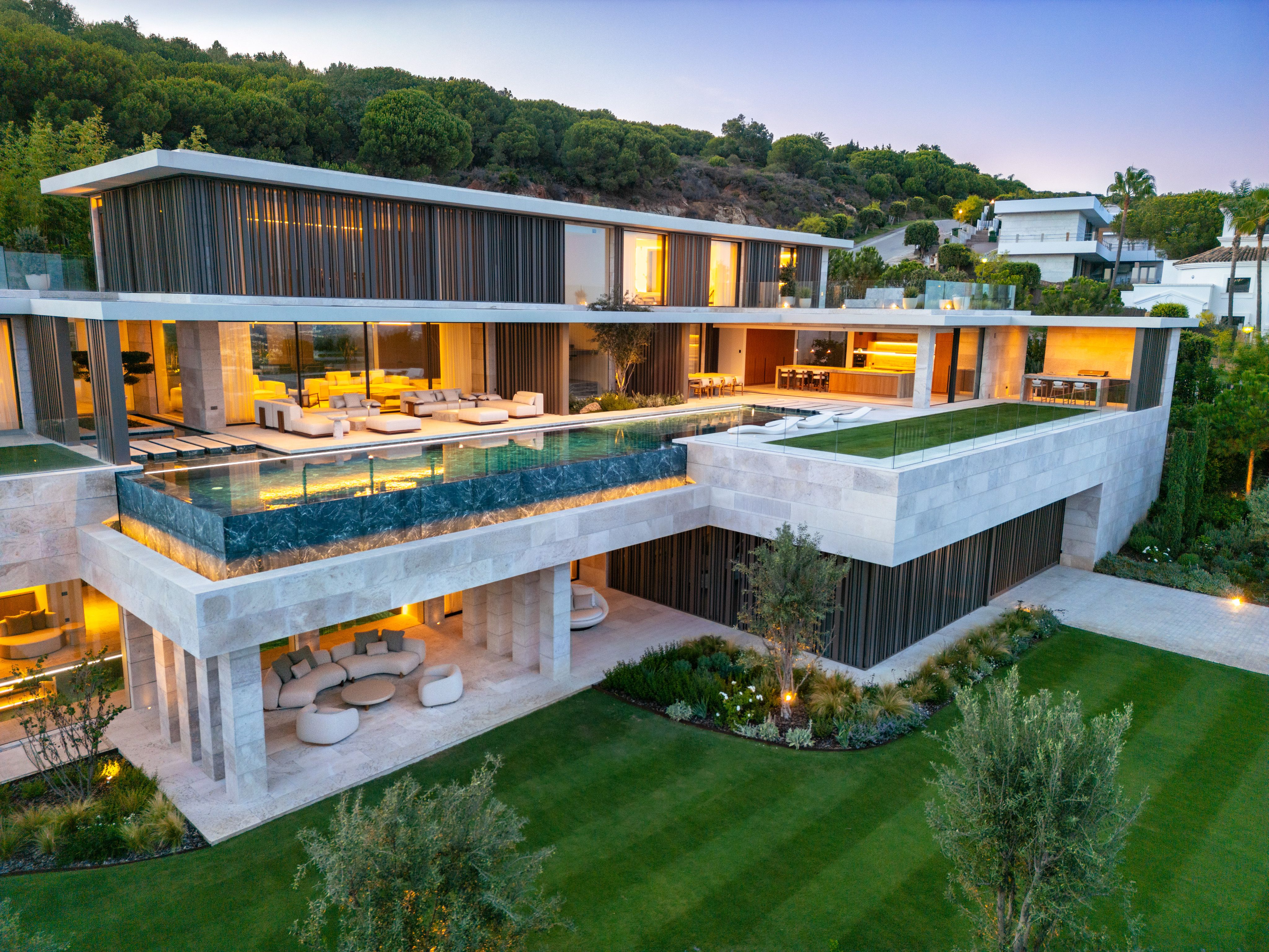 Aerial view at dusk of a contemporary stone and glass villa with a lit infinity pool, layered terraces, lawn and olive trees