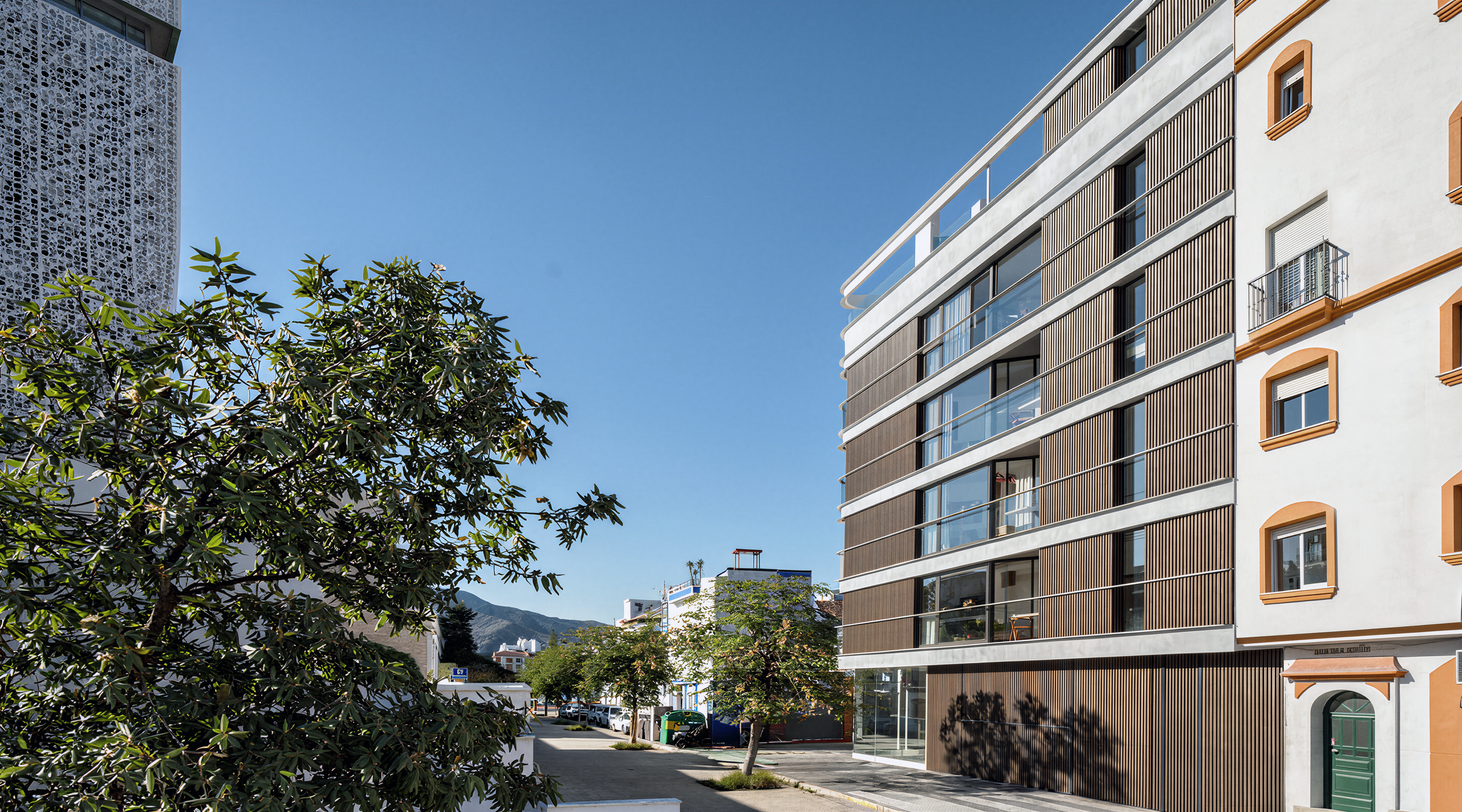 Computer-generated street view of a contemporary white apartment building with timber-look slatted screens and glass balconies beside a traditional Andalusian townhouse, mountains beyond