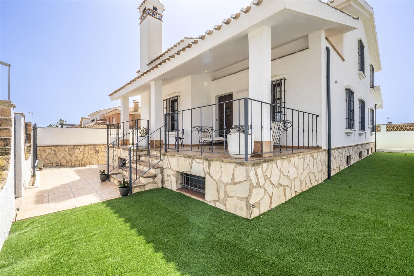 White villa with covered terrace, stone-clad base and a green garden in bright daylight.
