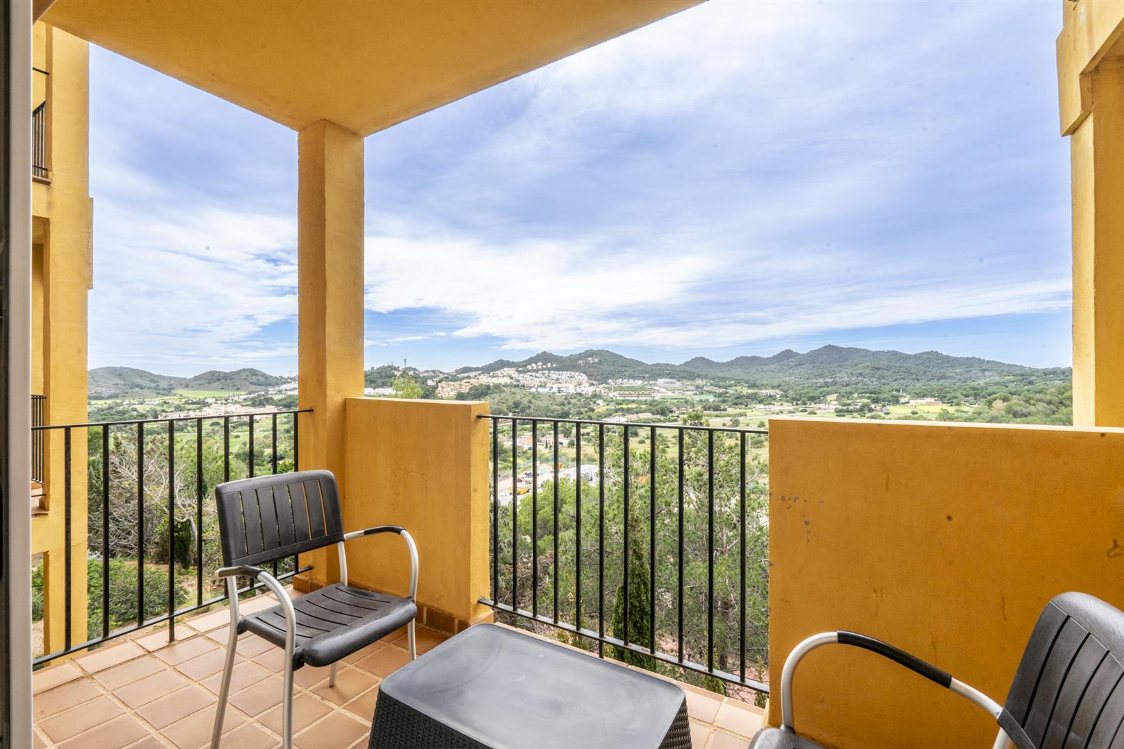 Covered ochre-walled balcony with black railing, two chairs and a low table looking over green hills, a white hillside village and mountains.