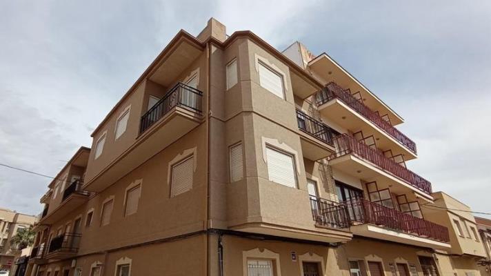 Photograph of a corner apartment building with a tan render facade, balconies and white window surrounds, seen from street level