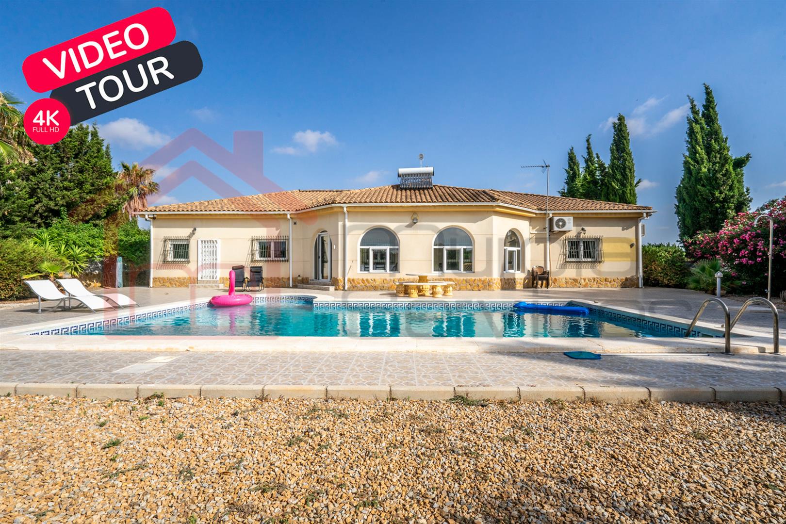 Photograph of the single-storey villa seen across the swimming pool, with sun loungers on the terrace and cypress trees behind