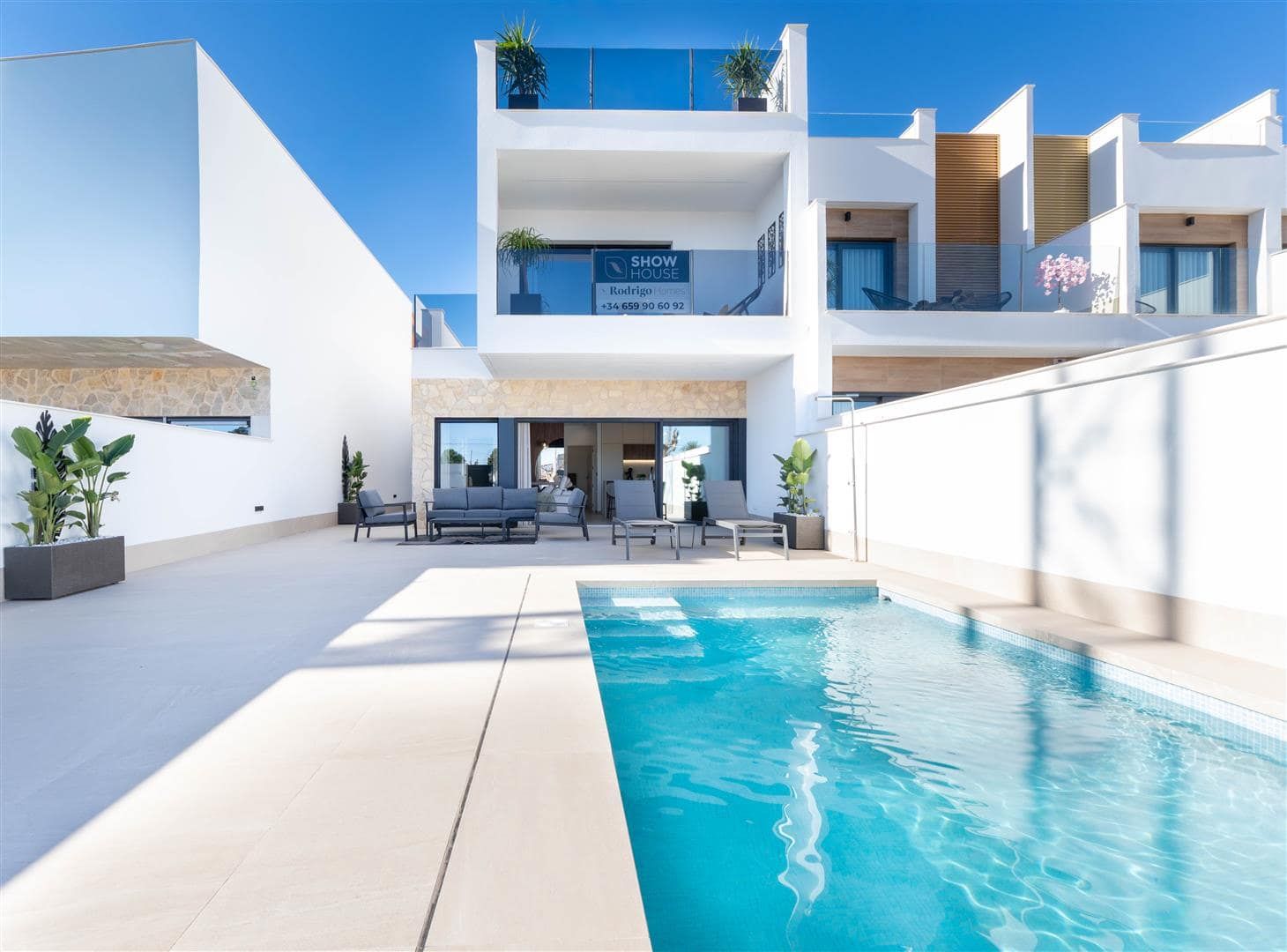 Front-facing day exterior view across a swimming pool towards a white two-storey home and terrace