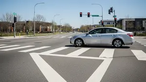 A wide city street with a white painted crosswalk and a silver sedan stopped just before the lines under a clear blue sky.