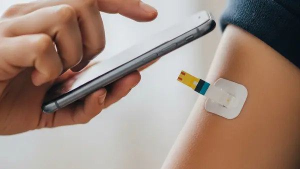 A close-up of a continuous glucose monitor sensor applied to a person's upper arm, while they hold a smartphone to scan the device for a blood sugar reading.
