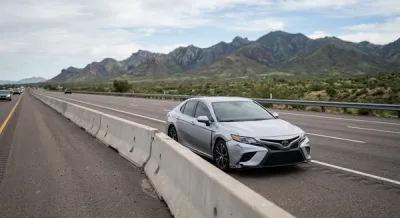 A view of a modern silver sedan on a highway shoulder next to a traffic divider with mountains in the background, symbolizing a roadside accident scene.