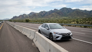 A view of a modern silver sedan on a highway shoulder next to a traffic divider with mountains in the background, symbolizing a roadside accident scene.