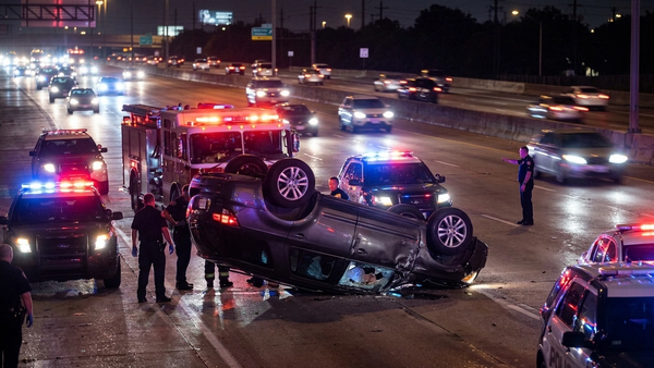 An overturned SUV rests on its roof on a dark Houston highway, surrounded by emergency response vehicles with flashing blue and red lights.