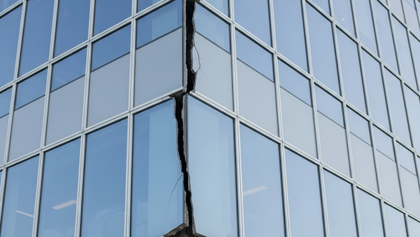 A modern glass building with a visible structural crack running down its side while a person in a safety vest and helmet inspects the foundation.