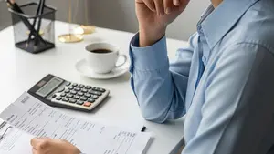 A person in professional attire looking thoughtfully at a paycheck while sitting at a desk with a calculator and a cup of coffee nearby.
