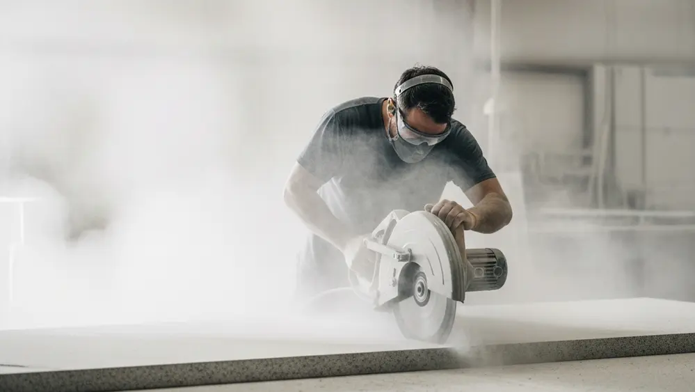 A stone fabrication worker in a mask cutting a slab of granite with a circular saw, surrounded by a cloud of fine white dust in a large industrial workshop.
