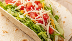 A close-up of a fresh flour tortilla taco filled with shredded green lettuce, diced tomatoes, and cheese, resting on a brown parchment paper sheet.