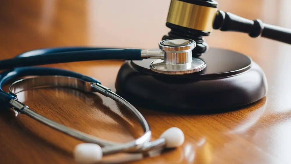 A close-up of a stethoscope lying on a wooden table next to a gavel, symbolizing the intersection of medicine and the law.