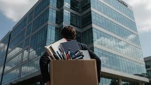 A person carrying a cardboard box filled with office supplies walks away from a sleek, multi-story glass insurance brokerage building during a bright afternoon.