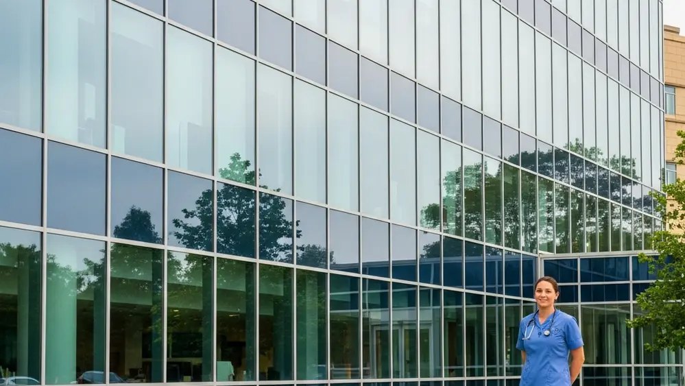 A modern hospital exterior with glass windows and a nurse in blue scrubs standing in the foreground near a green lawn.