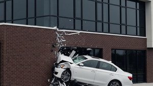 A white sedan with front-end damage is lodged into the side of a modern brick and glass building in a suburban setting.