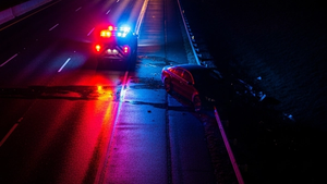 An aerial view of a roadside collision scene at night with emergency vehicle lights reflecting off a dark sedan near a highway shoulder.