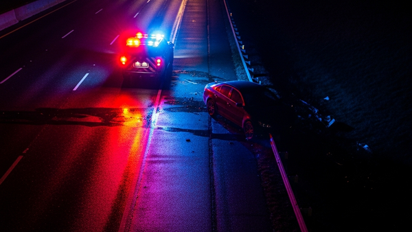 An aerial view of a roadside collision scene at night with emergency vehicle lights reflecting off a dark sedan near a highway shoulder.