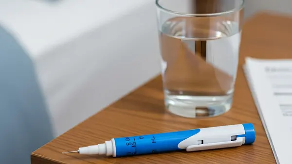 A close-up of a blue and white Dupixent injector pen lying on a wooden bedside table next to a glass of water, highlighting the medical device.